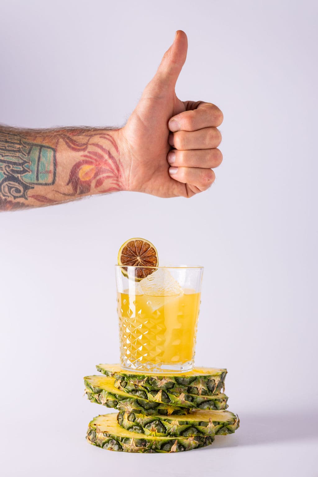 A tattooed hand giving a thumbs up above a yellow cocktail resting on pineapple slices