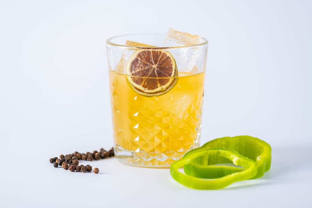 A yellow cocktail with a dried lime wheel beside peppercorns and green bell pepper rings
